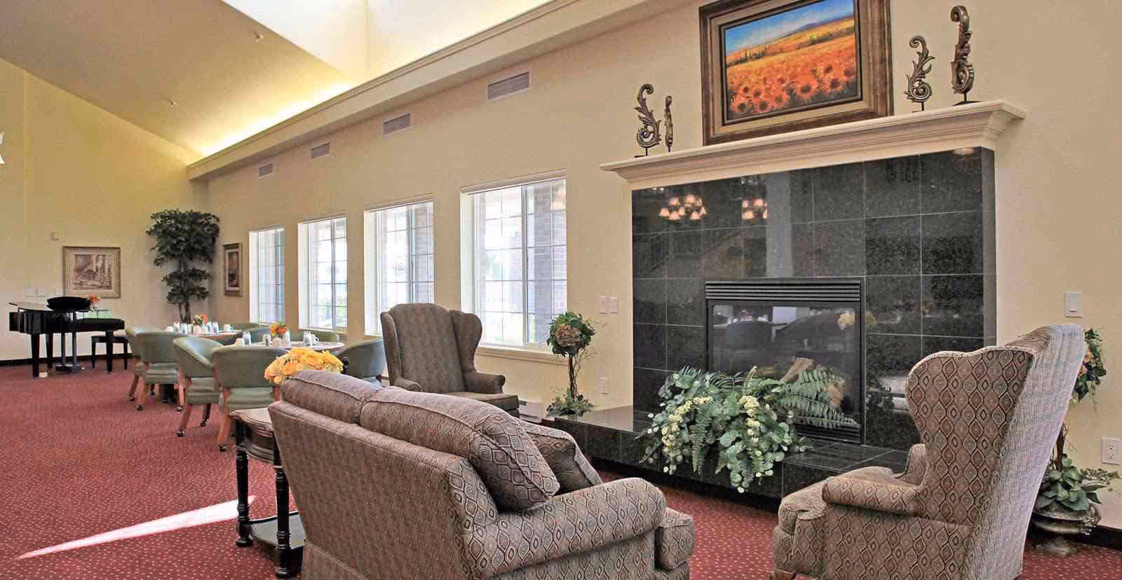 A cozy living room area with patterned upholstered armchairs and a sofa arranged around a black tiled fireplace decorated with greenery. The room has large windows letting in natural light, a red carpeted floor, and a grand piano in the background near a dining area with tables and chairs.