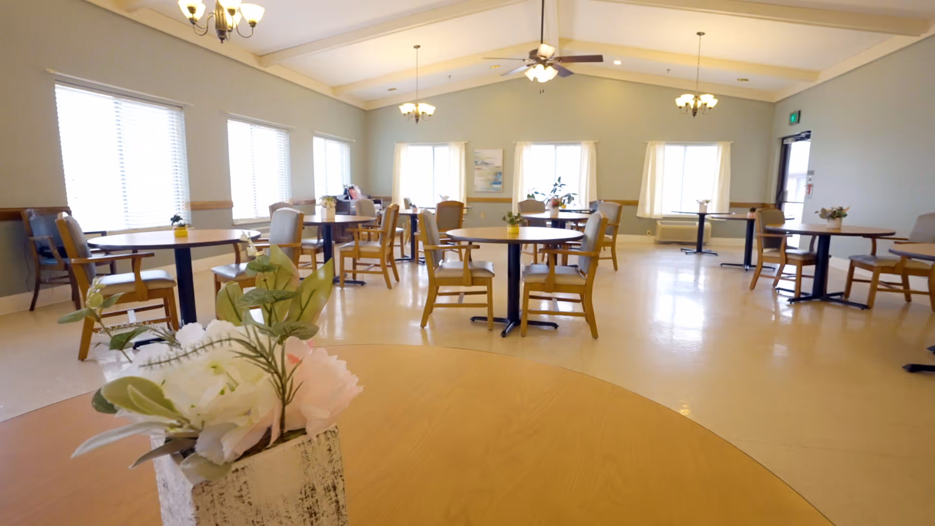 A spacious dining room with multiple round tables and wooden chairs arranged neatly. Each table has a small floral centerpiece. The room has large windows with blinds and curtains, light-colored walls, and ceiling fans with lights. The floor is light-colored and the room is well-lit with natural and artificial light.