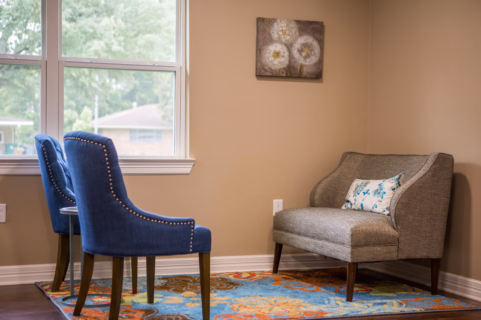 A cozy seating corner with two blue upholstered chairs by a window and a gray upholstered loveseat on a colorful rug.