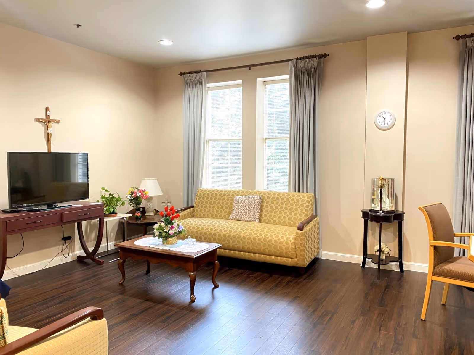 Sunlit common living room with a patterned yellow sofa, wooden coffee table with flowers, a TV on a console, and windows with curtains.