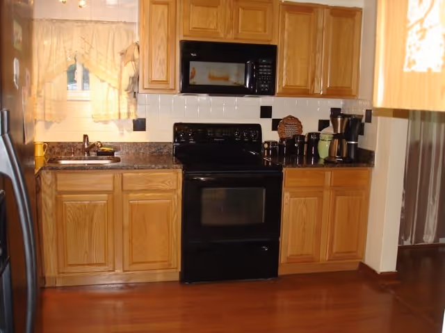 A kitchen with wooden cabinets, a black stove and microwave, a coffee maker, and a sink under a window with white curtains. The countertop is dark with a speckled pattern, and the floor is wooden.