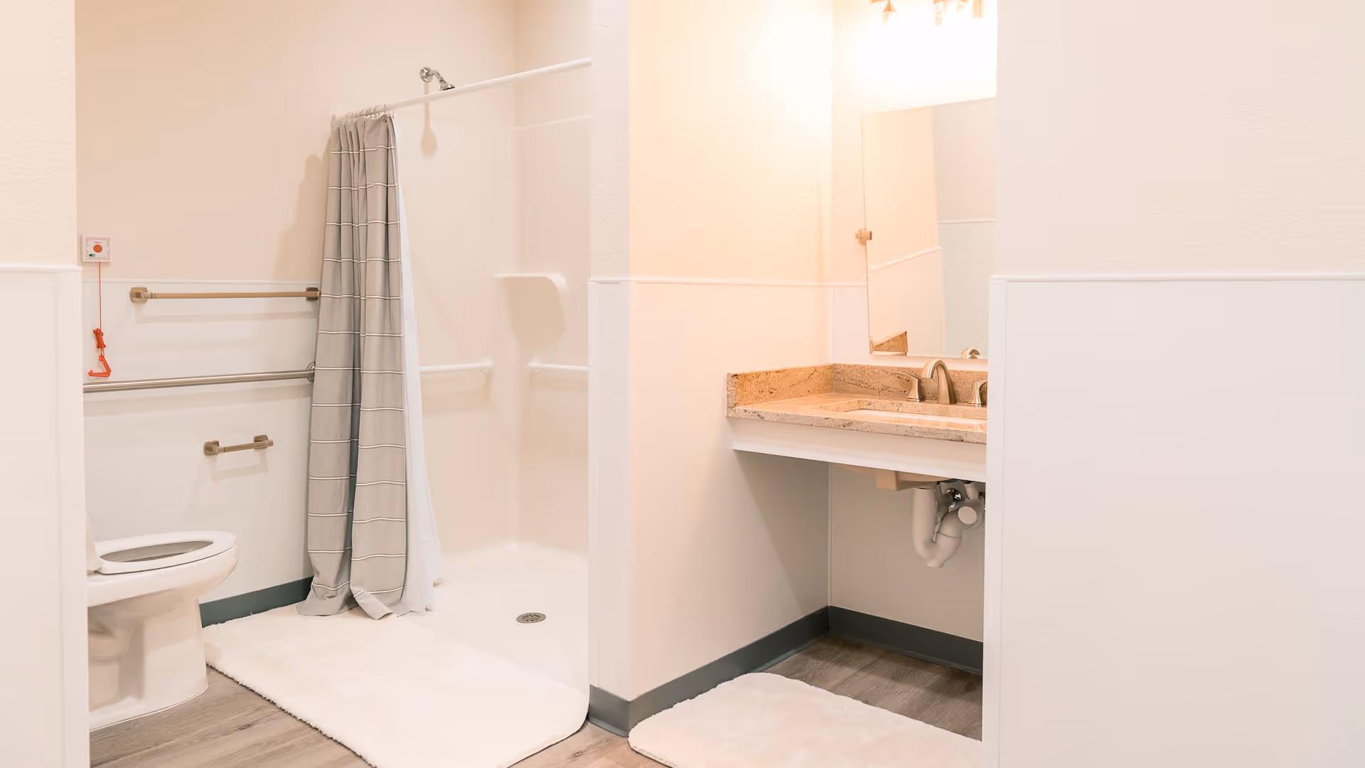 A clean and accessible bathroom featuring a toilet with grab bars, a walk-in shower with a curtain, and a vanity with a sink and mirror. The floor has a light wood finish and white bath mats are placed in front of the shower and sink.