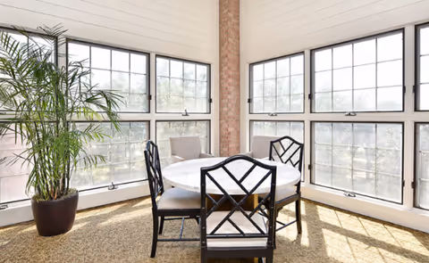 A bright corner dining area with large windows on two sides allowing natural light to fill the space. The room features a round white table surrounded by four black chairs with cushioned seats. A tall potted plant is placed near the windows, and the floor is carpeted.