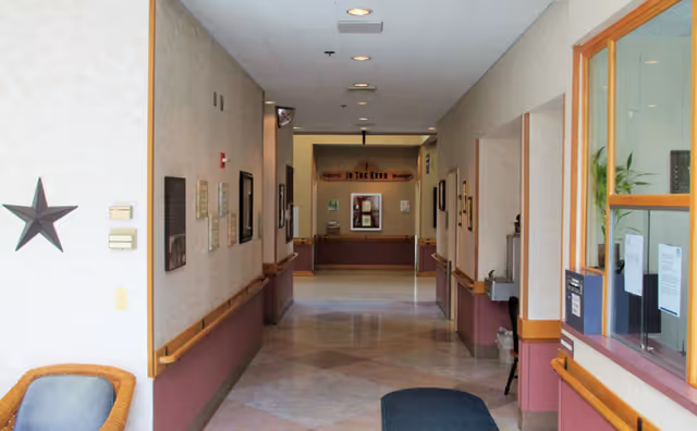 A clean, well-lit hallway in a nursing and rehabilitation center with beige walls, handrails on both sides, framed pictures on the walls, a star decoration on the left wall, and a reception window on the right. The floor is tiled, and there is a blue mat near the entrance.