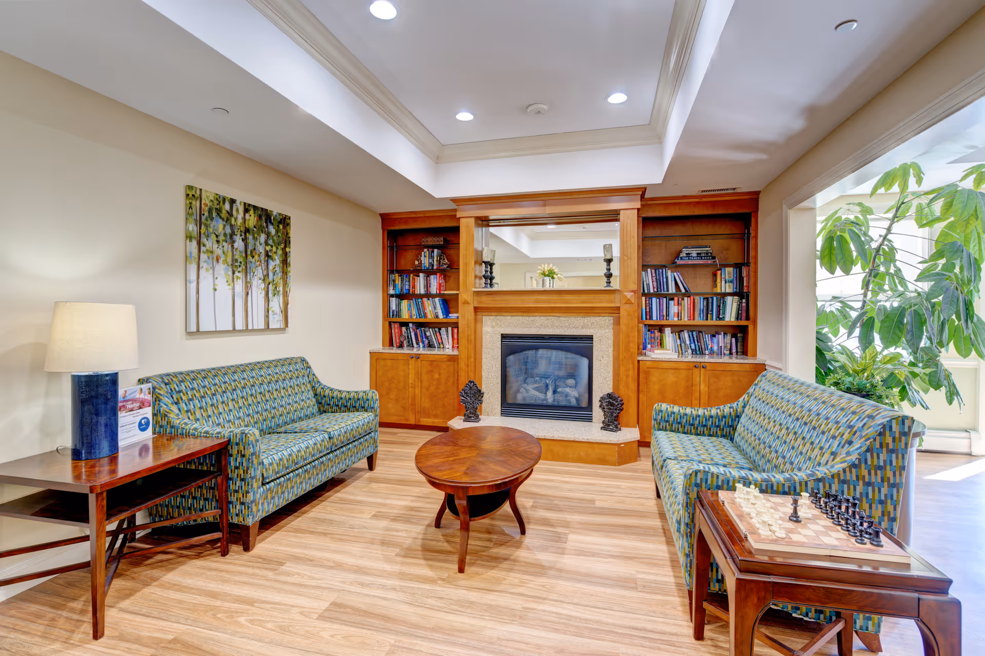 A cozy living room area with two patterned sofas facing each other, a wooden coffee table in the center, and a fireplace with built-in wooden bookshelves on either side. There is a lamp on a side table next to one sofa, a chessboard on a side table next to the other sofa, and a large plant near a window letting in natural light.