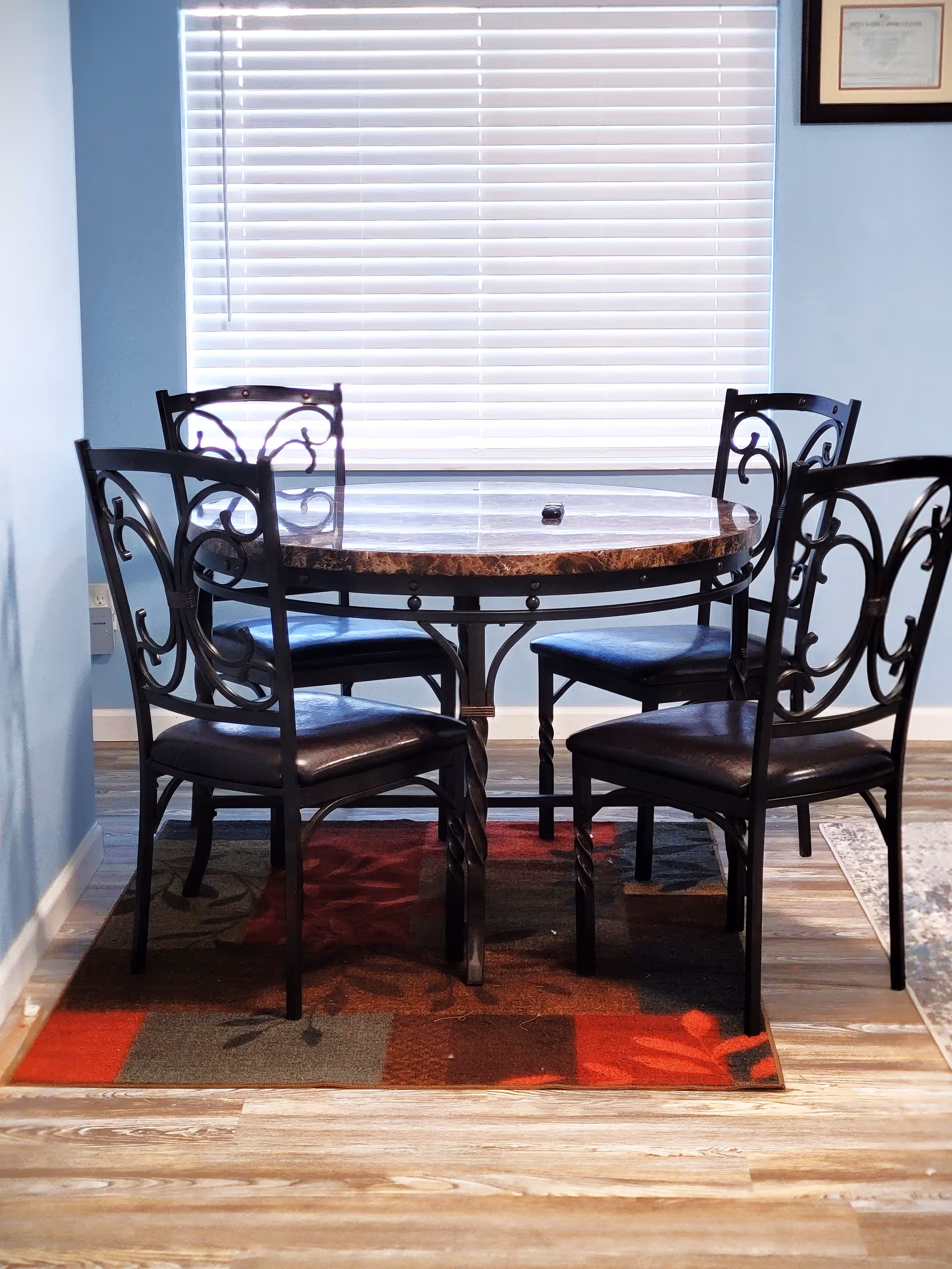 A round dining table with a marble-like top and four black metal chairs with decorative backs, placed on a multicolored rug with red, brown, and gray tones. The table is positioned in front of a window with closed white blinds, and the walls are painted light blue. A framed certificate is partially visible on the right wall.