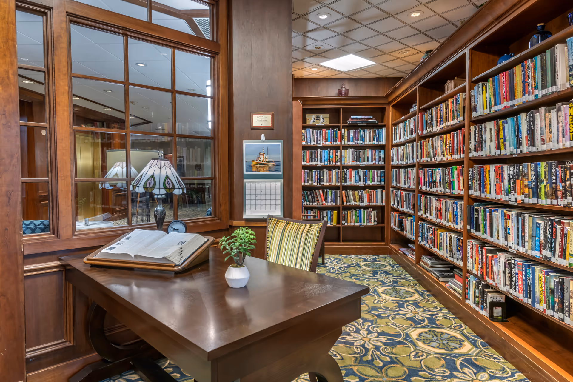 A cozy library room with wooden bookshelves filled with books lining the walls. A wooden desk with an open book, a small potted plant, and a decorative lamp sits in the foreground. The room has a patterned carpet and a window with wooden framing looking into another room.