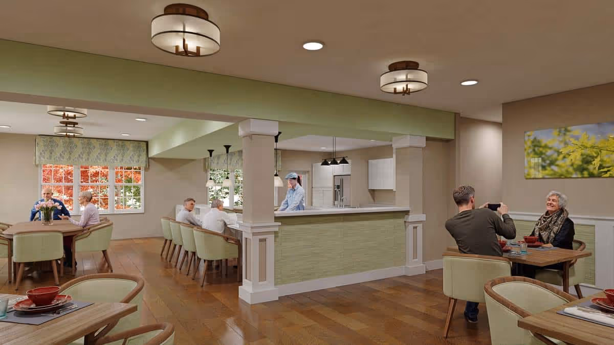 A bright dining room in The Timberley facility with several elderly people seated at tables enjoying meals. The room features wooden floors, green cushioned chairs, large windows with patterned valances, and a central kitchen area with a person preparing food. Modern ceiling lights illuminate the space, and a nature-themed painting hangs on the wall.