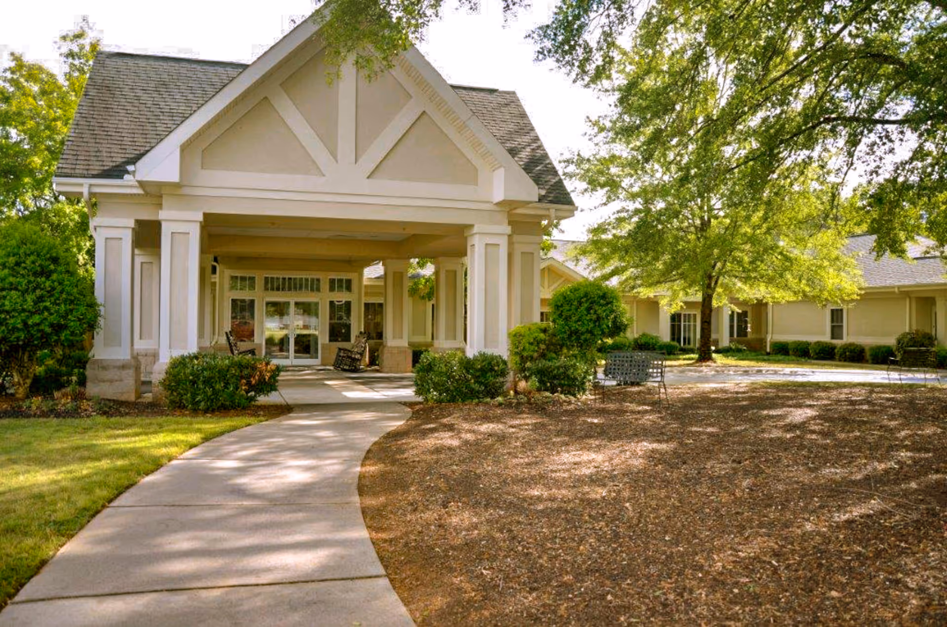 Front entrance of a senior living facility with a covered porte-cochere, walkway, benches, and landscaped trees and shrubs.