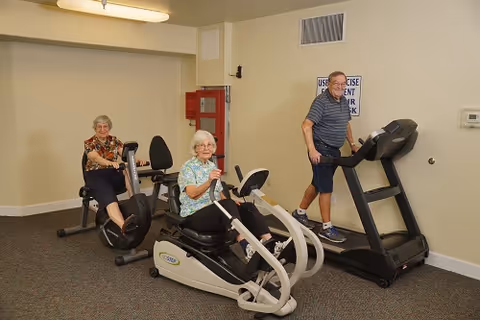 Three elderly individuals exercising in a fitness room. Two women are seated on recumbent exercise bikes, and a man is walking on a treadmill. The room has beige walls, a carpeted floor, a fire extinguisher mounted on the wall, and a sign with exercise equipment usage instructions.
