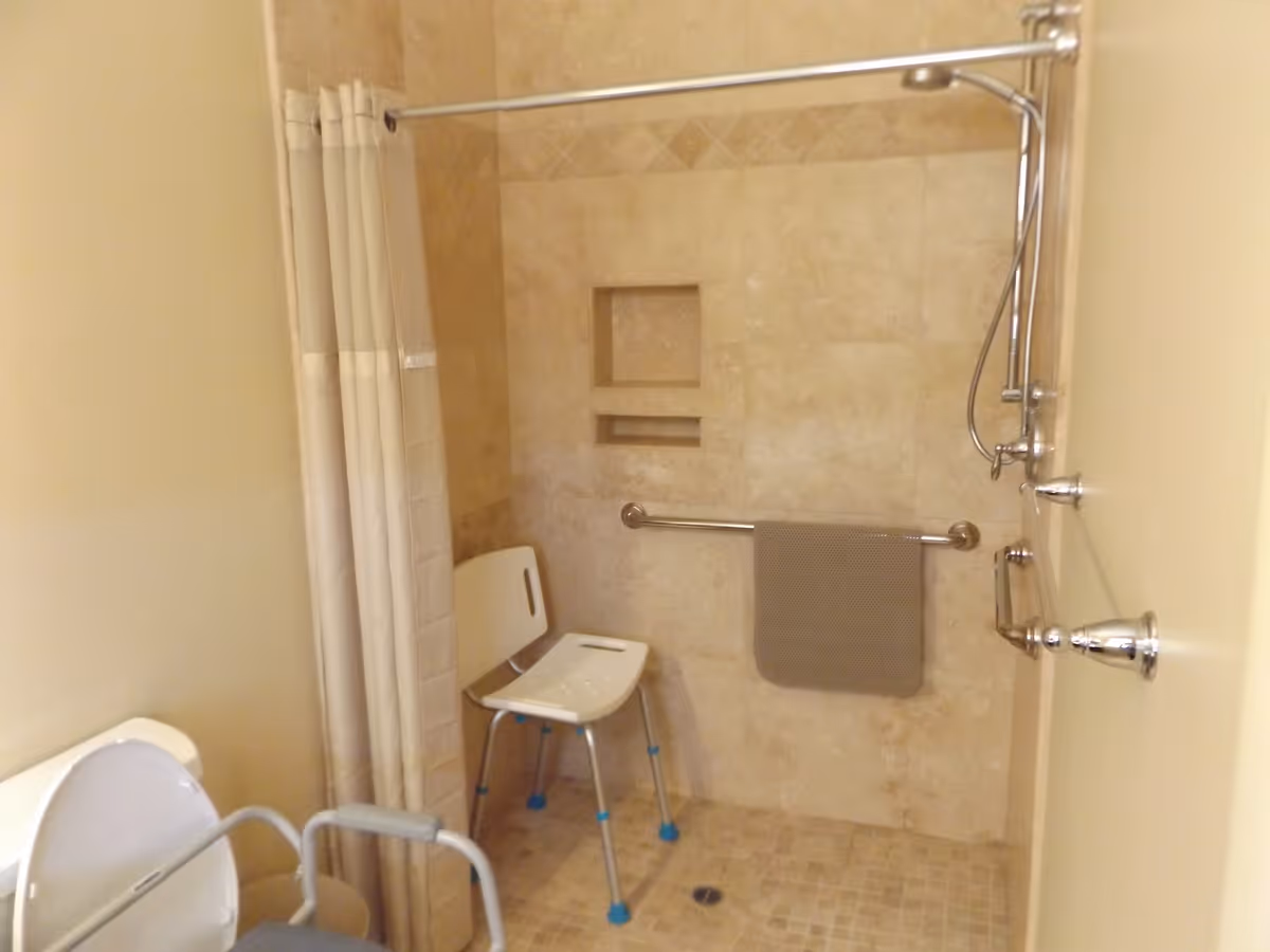 Accessible bathroom shower area with beige tiled walls and floor, a white shower chair with blue feet, a grab bar with a gray towel hanging on it, a handheld showerhead, and a shower curtain partially drawn to the side. A toilet with arm supports is partially visible on the left.
