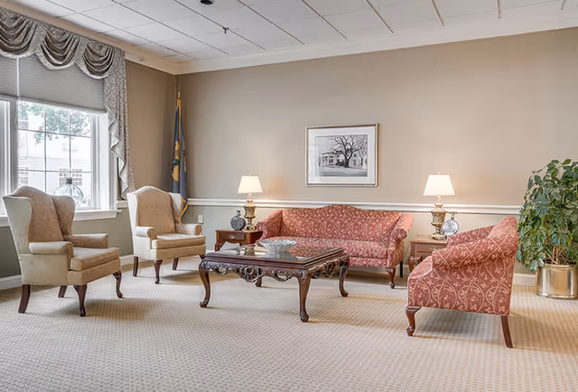 A bright communal living room with upholstered armchairs and matching sofas arranged around a carved wooden coffee table under lamps and framed art.