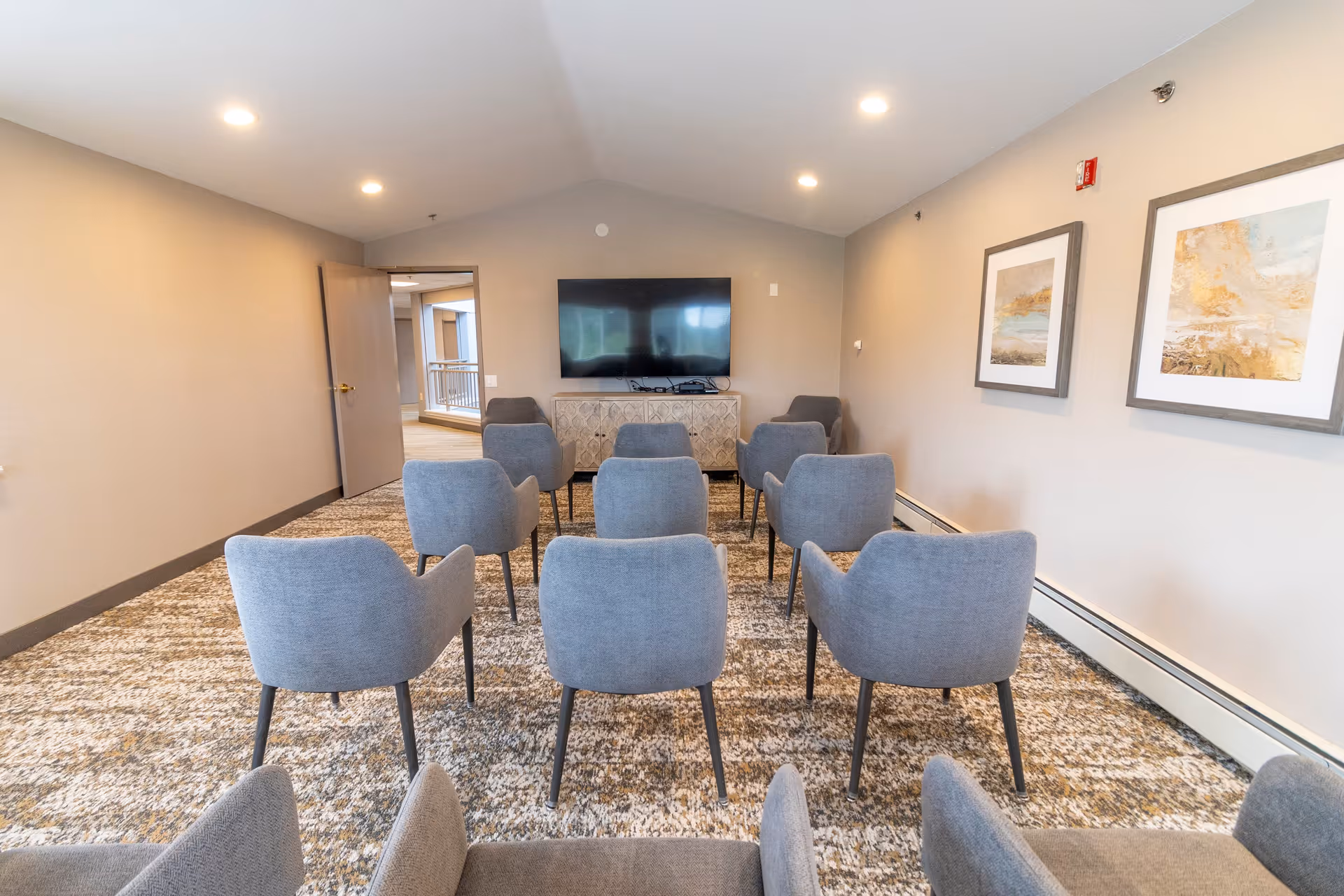 A small meeting or media room with two rows of gray upholstered chairs facing a flat-screen TV mounted on the wall. The room has beige walls, a patterned carpet, two framed abstract paintings on the right wall, and recessed ceiling lights. An open door at the back leads to a hallway.