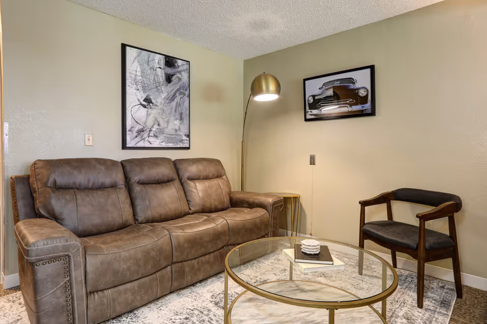 A cozy living room with a brown leather sofa, a wooden armchair with dark cushions, a round glass coffee table with books and decor on it, a tall gold floor lamp, and two framed artworks on the beige walls.