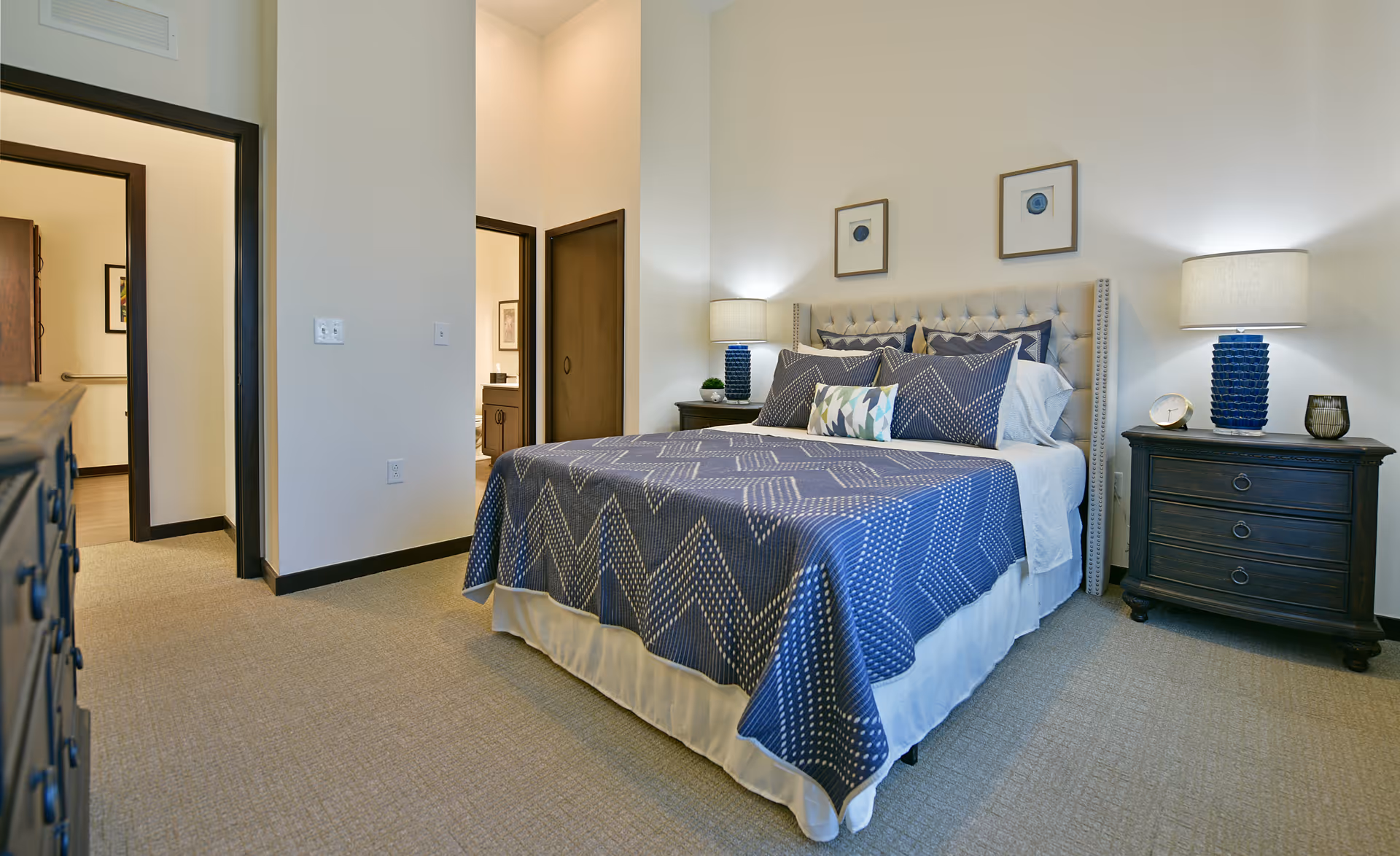 Well-lit bedroom with a tufted bed dressed in blue patterned bedding, two bedside tables with lamps, and an open doorway to adjacent rooms.