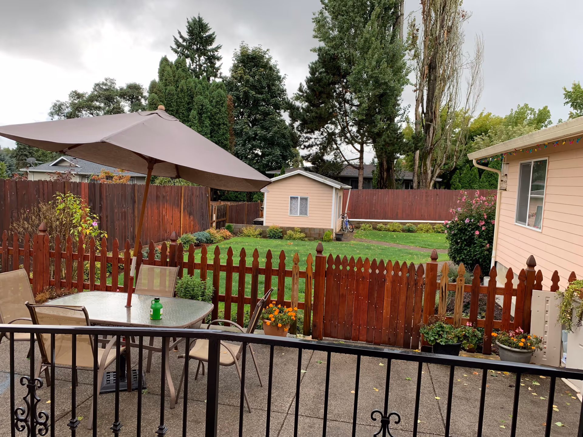 Outdoor patio area with a glass table and four chairs under a large beige umbrella. The patio is enclosed by a wooden picket fence with a gate leading to a green lawn and a small shed. There are various plants and flowers around the fence and the side of a peach-colored building is visible on the right. Tall trees and a cloudy sky are in the background.