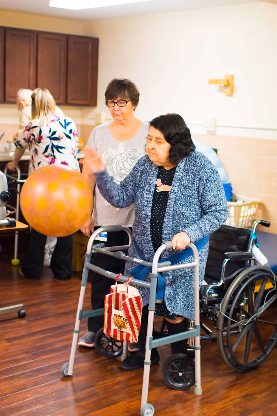 An elderly woman using a walker is playing with a large orange ball indoors, assisted by another woman standing behind her. In the background, other elderly individuals are engaged in activities near a kitchen area with wooden cabinets.