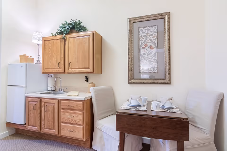 Small kitchenette with wooden cabinets, a sink and mini fridge beside a two-person dining table set under framed artwork.