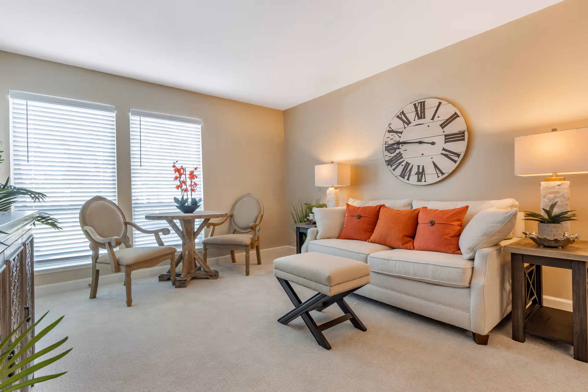 A cozy living room with a beige sofa adorned with three orange cushions, a small cushioned ottoman in front, two wooden chairs with beige upholstery around a small round wooden table with a red flower arrangement, two large windows with white blinds, a large round wall clock with Roman numerals above the sofa, and two side tables each with a lamp and decorative plants.