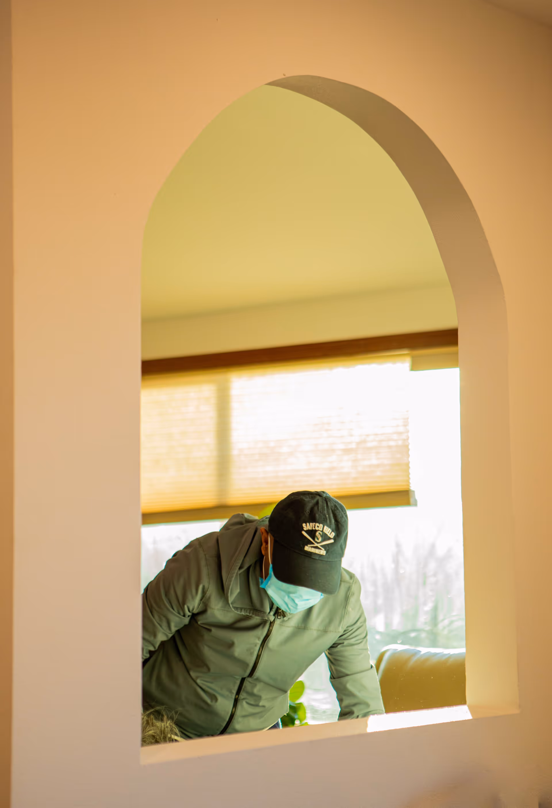 A person wearing a green jacket, a black cap with 'SAFECO FIELD' written on it, and a blue face mask is seen through an arched wall opening inside a room with a window covered by a beige blind.