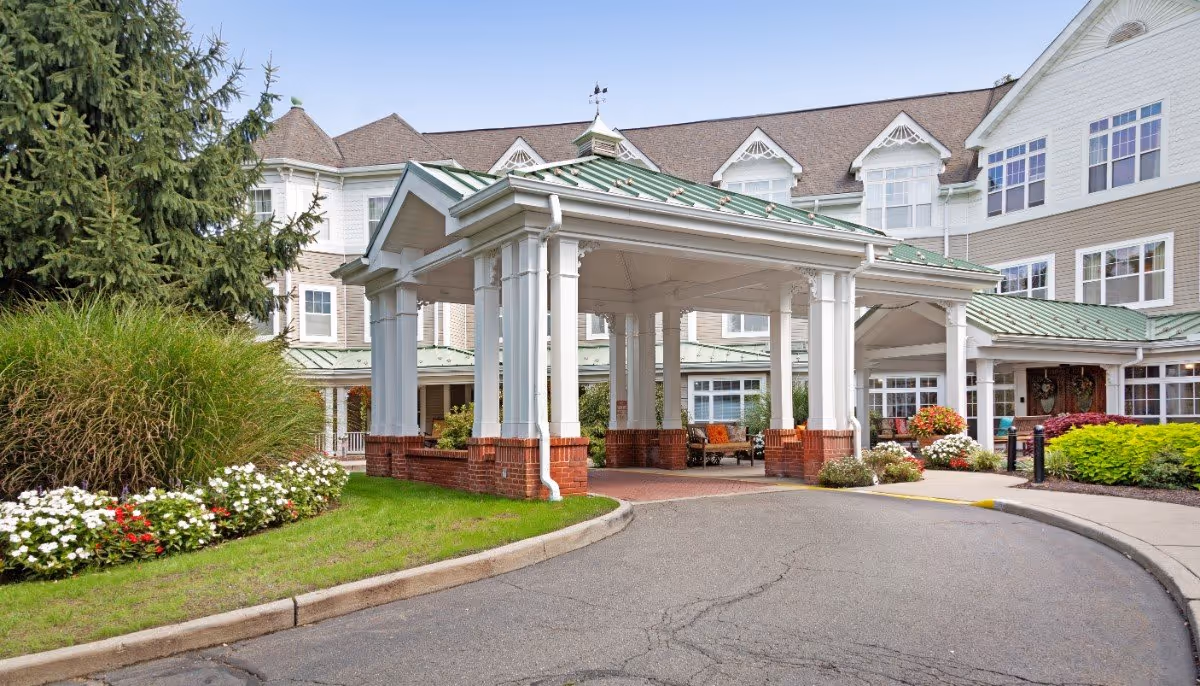 Exterior view of a senior living facility with a covered entrance supported by white columns with brick bases. The building has multiple windows and a green metal roof. There are landscaped flower beds with white and red flowers and green shrubs around the entrance area.