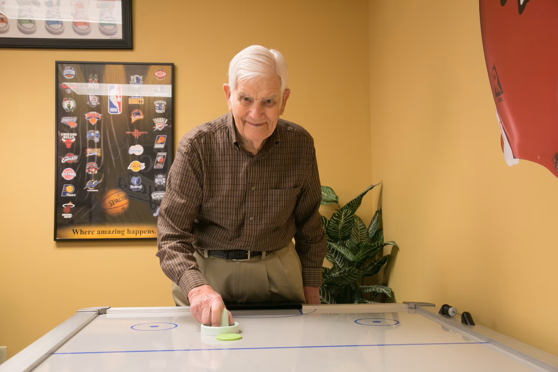 An elderly man with white hair wearing a brown checkered shirt and beige pants is playing air hockey indoors. Behind him, there is a framed poster featuring NBA team logos and a basketball with the text 'Where amazing happens.' A green leafy plant is placed in the corner of the room with yellow walls.