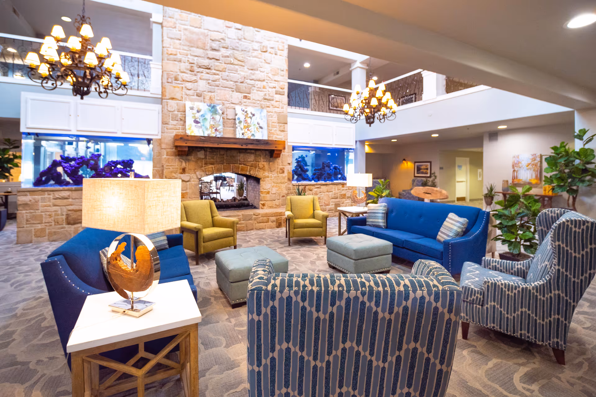Spacious furnished lounge with blue and patterned seating arranged around a stone fireplace, chandeliers, and plants in a senior living facility.