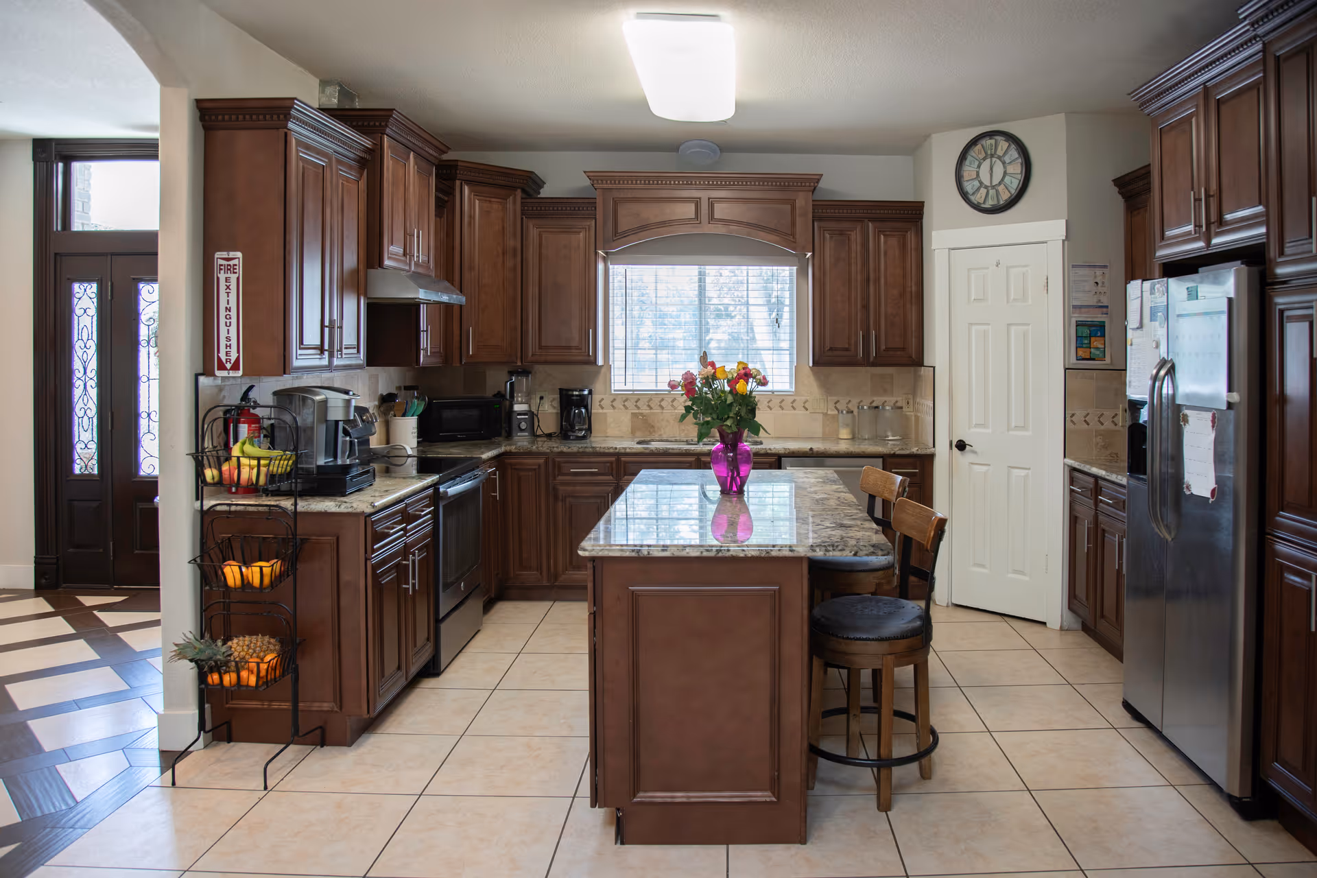 A spacious kitchen with dark wooden cabinets, a central island with a granite countertop, and two wooden bar stools. The kitchen features stainless steel appliances including a refrigerator and stove. A window above the sink lets in natural light, and a vase with colorful flowers is placed on the island. There is a fruit basket stand near the entrance, and a clock is mounted on the wall above a white door.
