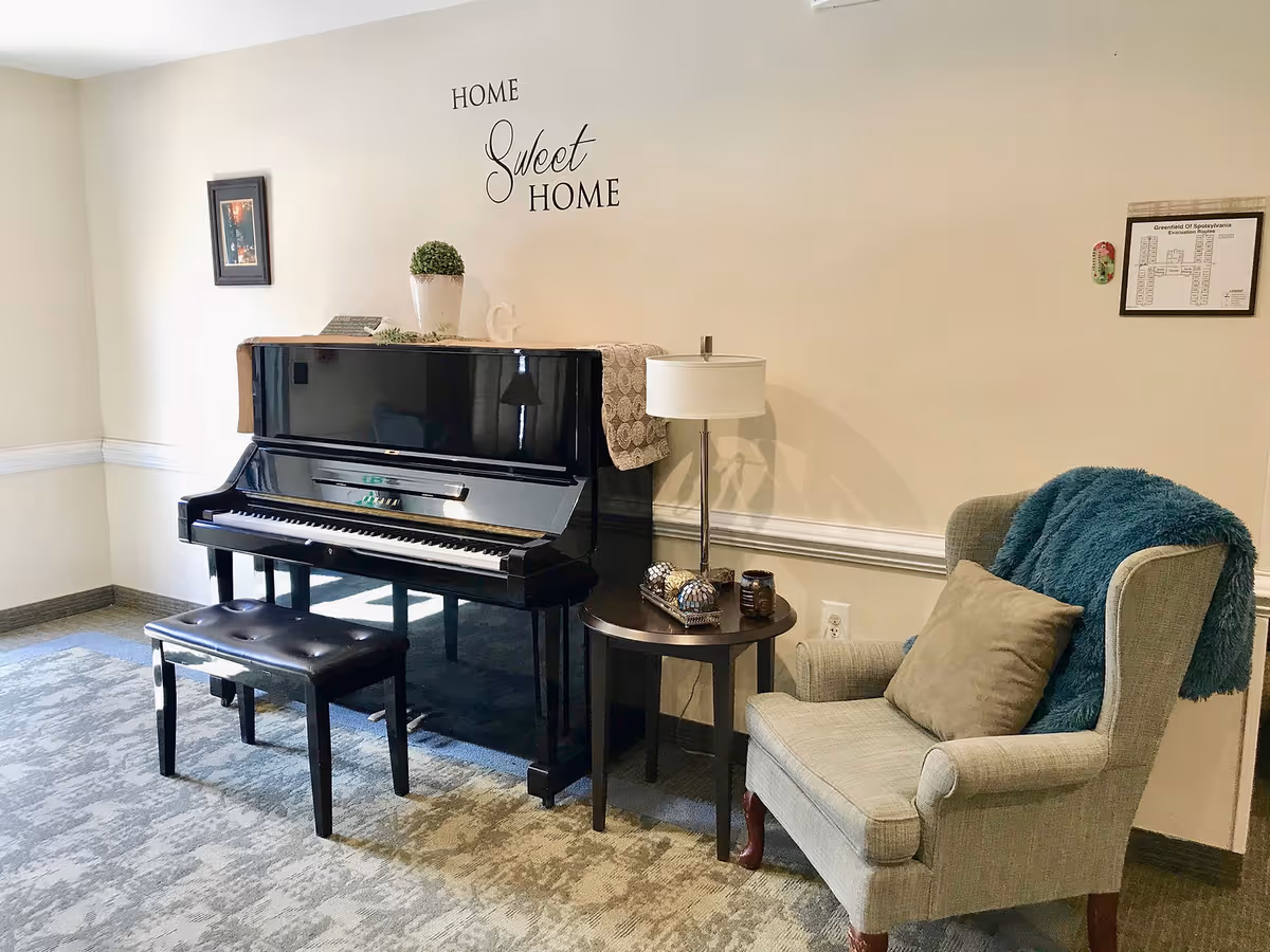 A cozy corner in a living area featuring a black upright piano with a matching bench, a small round side table with a lamp and decorative items, and a comfortable armchair with a pillow and a teal throw blanket. The wall behind has the phrase 'Home Sweet Home' and two framed pictures.