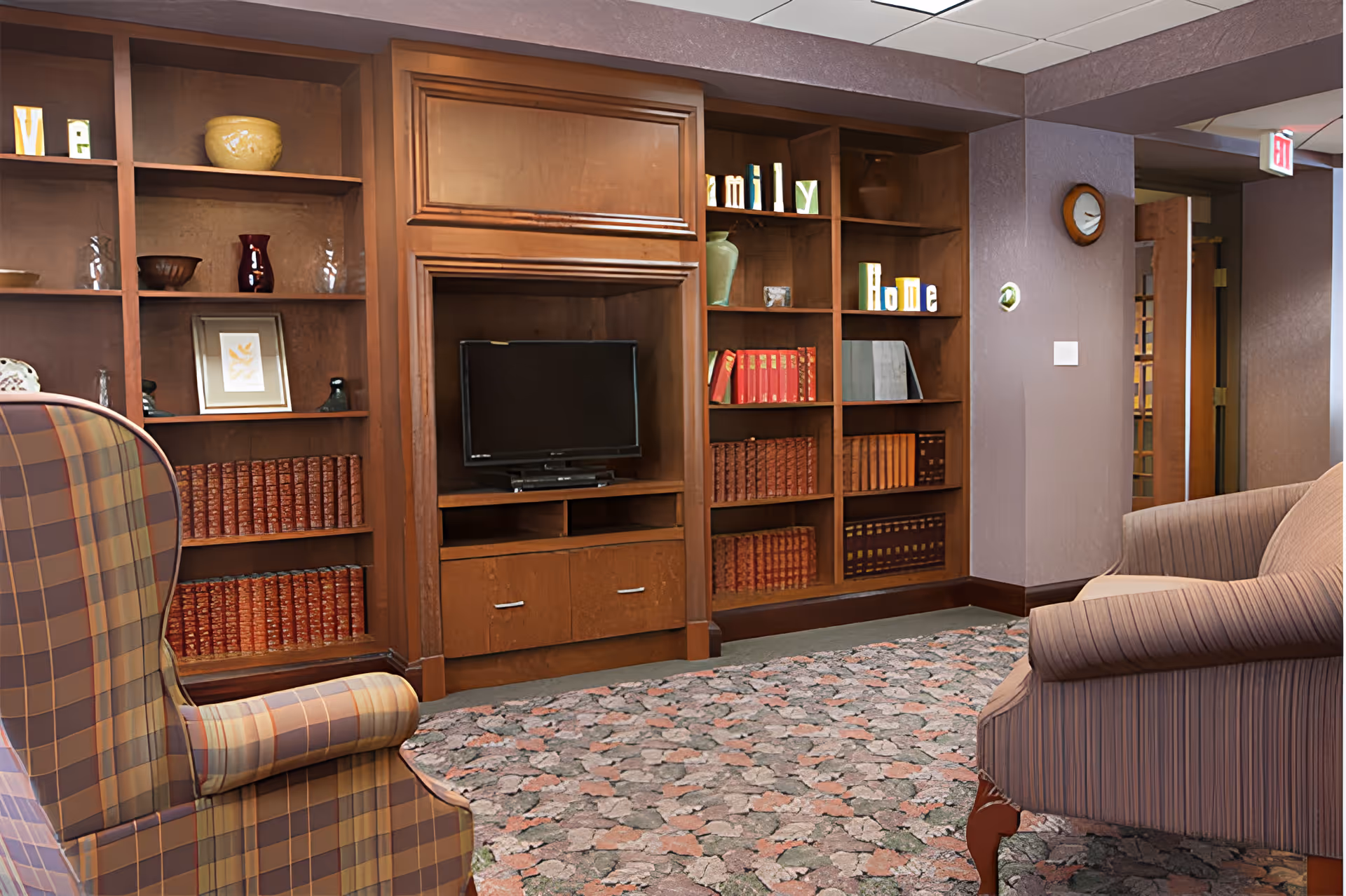 A cozy living room area with a patterned armchair and a striped armchair facing a wooden entertainment center with a flat-screen TV. The entertainment center is flanked by built-in wooden bookshelves filled with books, decorative items, and blocks spelling out 'Family' and 'Home'. The room has a floral carpet and purple walls with a clock and an exit sign visible in the background.