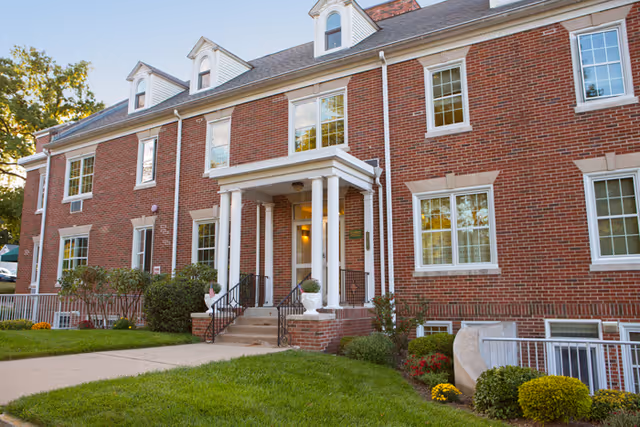 Exterior view of a two-story red brick building with white-framed windows and a small covered entrance supported by white columns. The building is surrounded by green grass, bushes, and flowers, with a concrete walkway leading to the entrance.