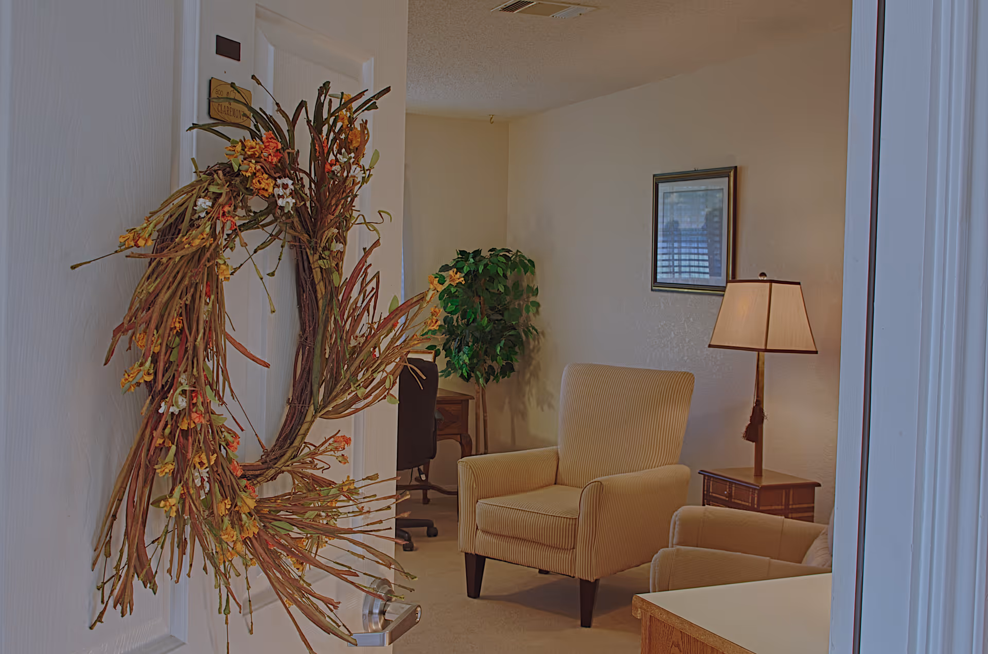 View into a cozy living area with a beige armchair, a side table with a lamp, a framed picture on the wall, and a potted plant in the corner. A door with a decorative wreath is partially open in the foreground.