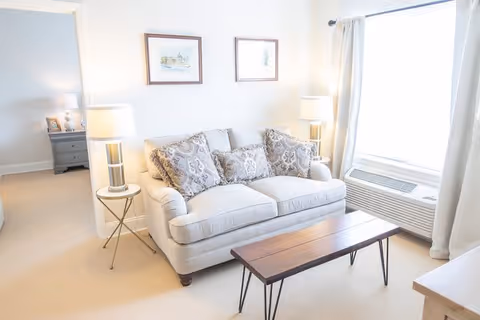 A bright living room with a beige sofa adorned with patterned cushions, a wooden coffee table with metal legs in front of it, two side tables each with a lamp, framed artwork on the wall above the sofa, a large window with white curtains, and an air conditioning unit below the window.