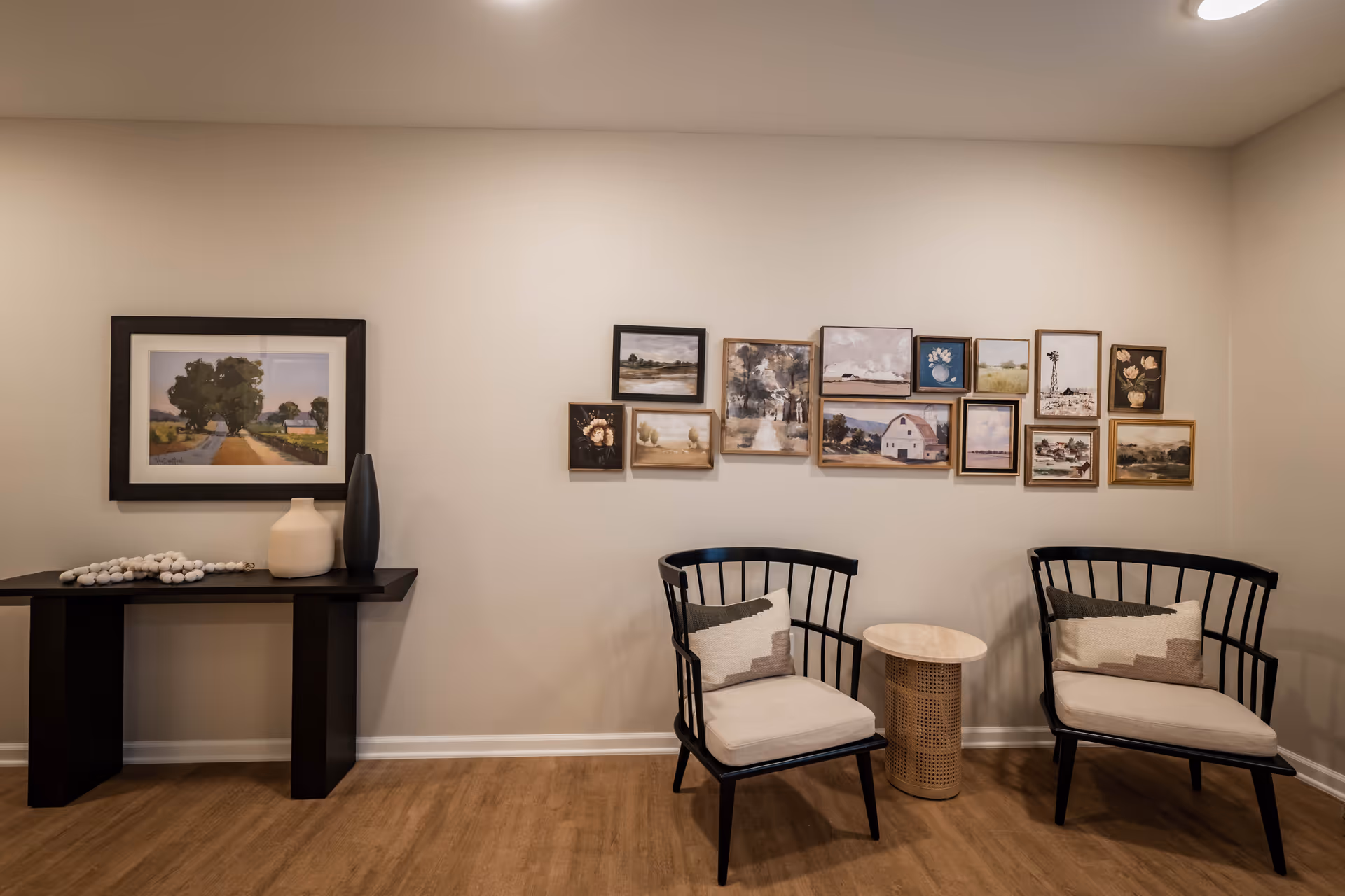A cozy seating area with two black wooden chairs with beige cushions and patterned pillows, separated by a small round wicker side table. On the wall behind the chairs is a gallery of framed paintings depicting landscapes and floral scenes. To the left, there is a black console table with decorative vases and a string of white beads, and a framed landscape painting hanging above it. The room has light beige walls and wood flooring.
