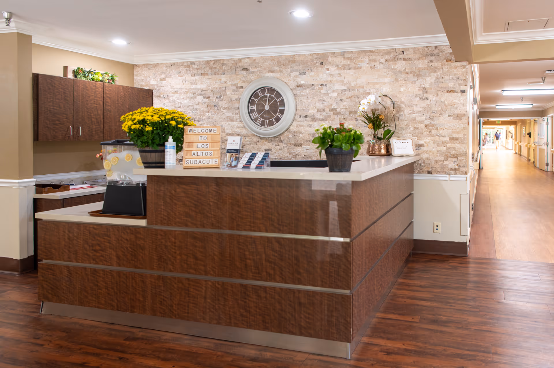 Reception desk area in a senior living facility with a wooden counter, a large wall clock, potted plants, a water dispenser with lemon slices, and a sign that reads 'Welcome to Los Altos Subacute'. The background shows a hallway with wooden flooring and beige walls.