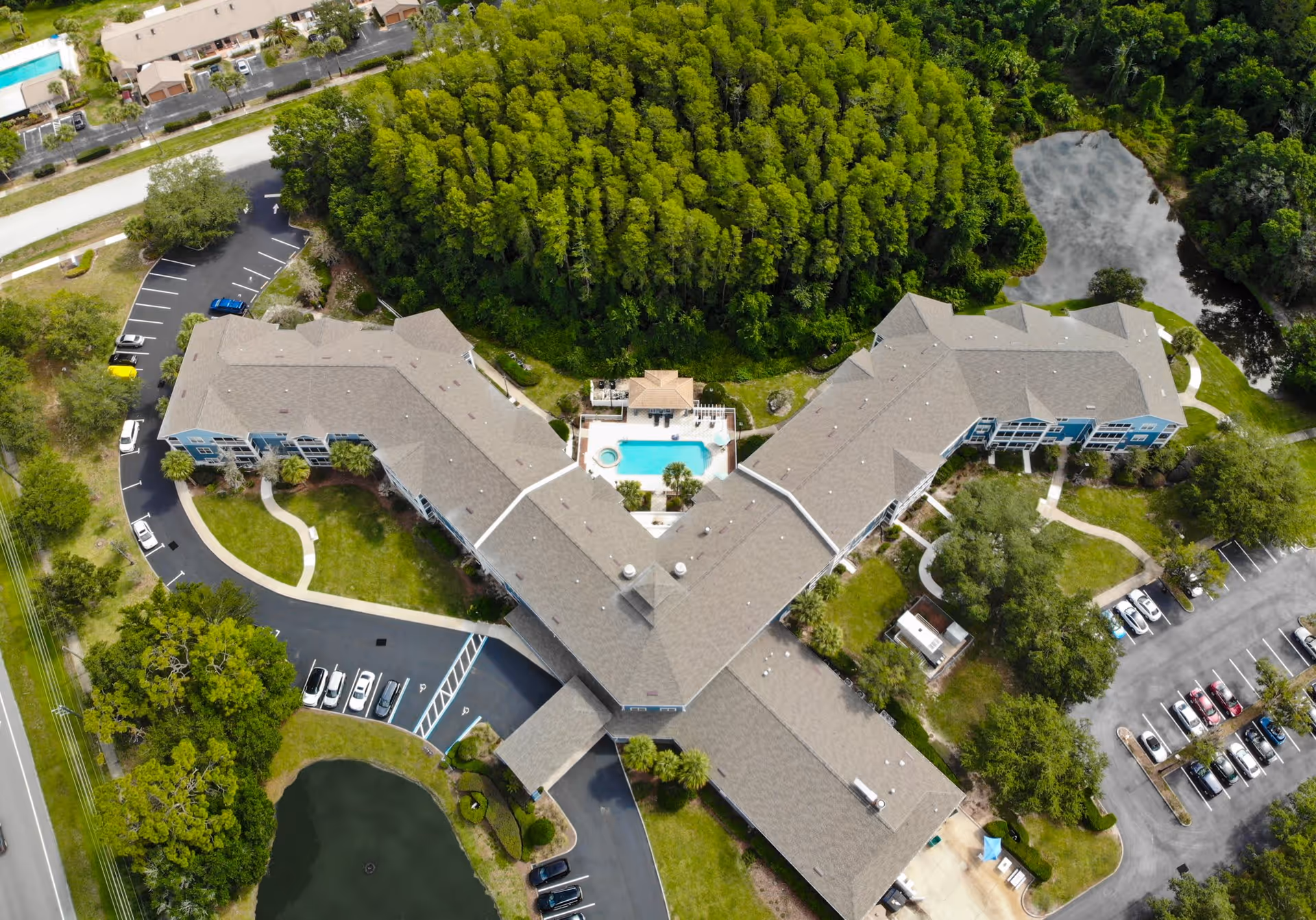 Aerial view of Addington Place of East Lake senior living facility showing a large building complex with a central swimming pool, surrounded by parking lots, green lawns, trees, and a small pond.