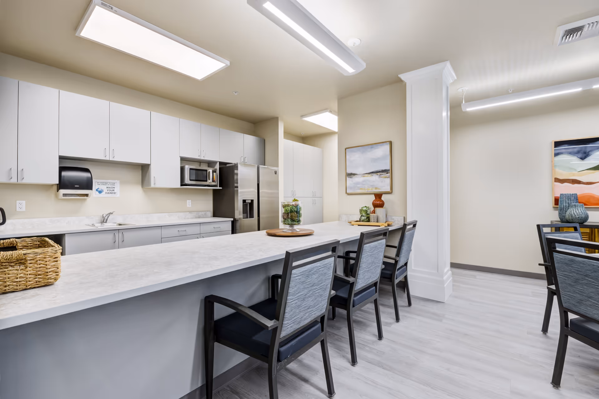 Bright communal kitchen and dining area with a long countertop island, seating, white cabinets, and stainless steel appliances.