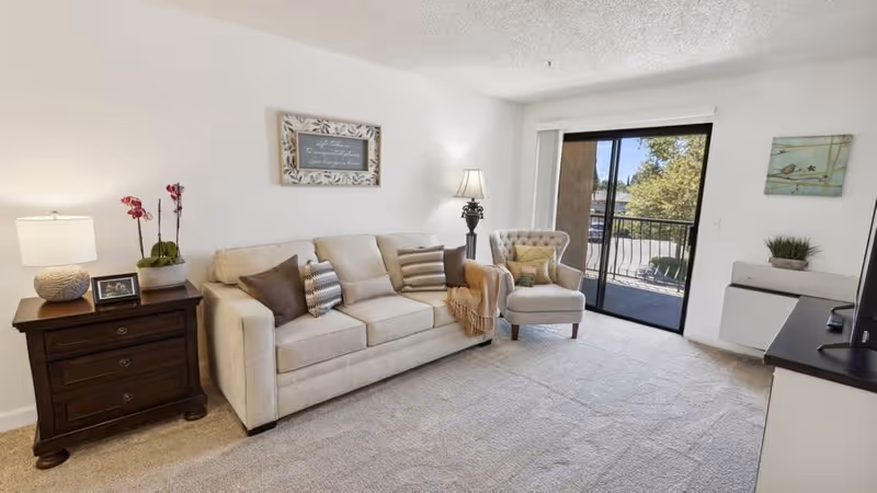 A bright living room with a beige sofa adorned with several pillows and a throw blanket, a cushioned armchair, a wooden side table with a lamp, a potted orchid, and a framed photo. There is a floor lamp next to the sofa, a sliding glass door leading to a balcony with a view of trees outside, and a small cabinet with a plant on top. The walls are white with two pieces of wall art.
