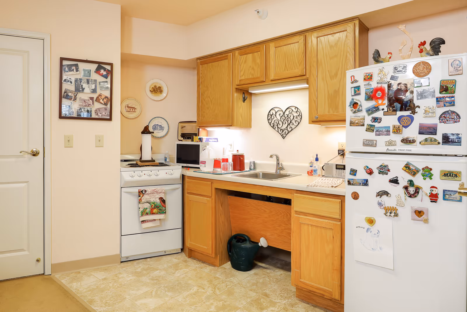 A small kitchen area with wooden cabinets, a white stove with a decorative towel hanging on the handle, a white refrigerator covered with various colorful magnets and drawings, a stainless steel sink with soap dispensers, and a watering can stored underneath the sink. The walls are decorated with framed photos and decorative plates, and there is a heart-shaped metal wall ornament above the sink.