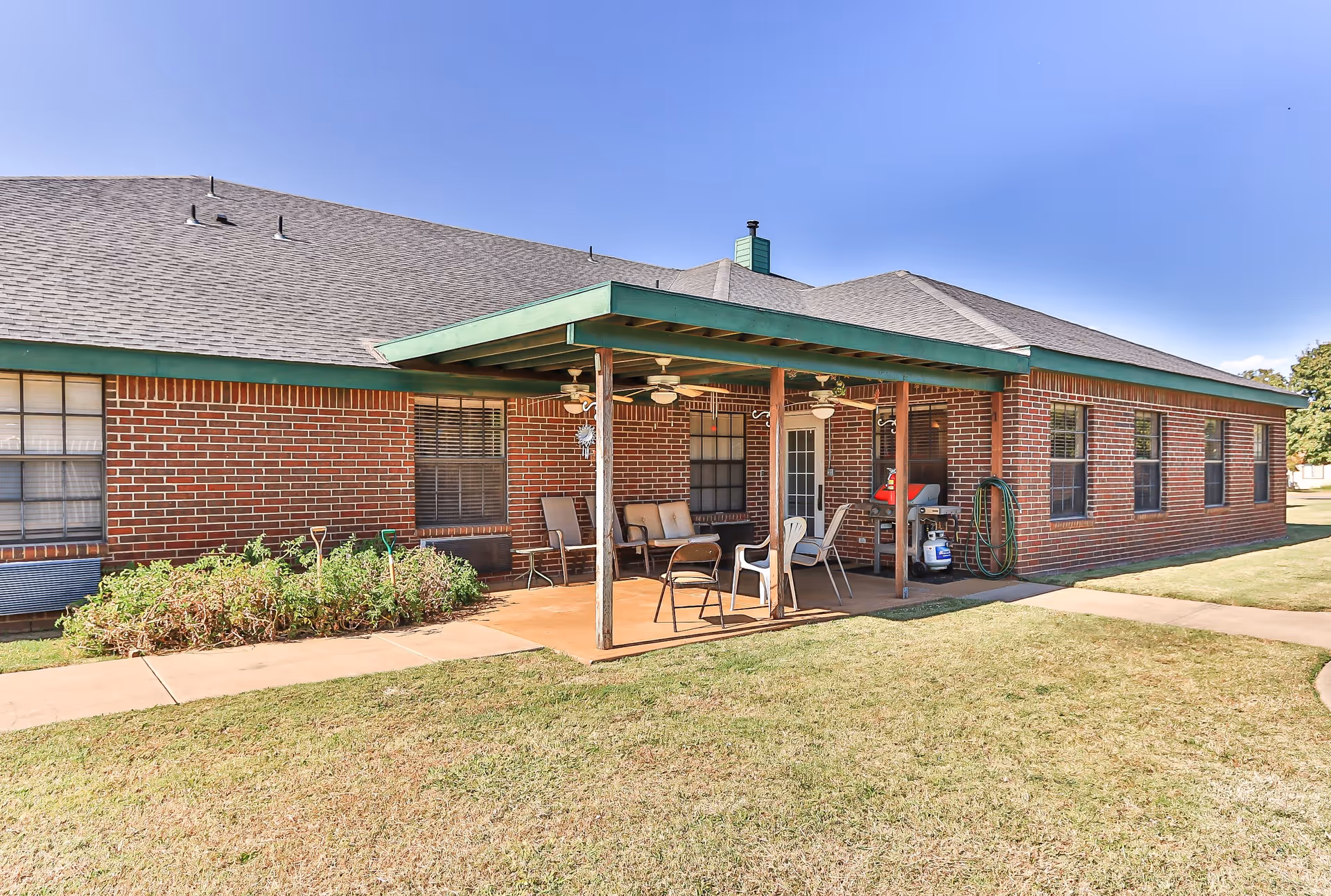 Outdoor patio area of a brick building with a green roof trim, featuring several chairs, a bench, ceiling fans, a grill, and garden tools near a small garden bed, under a clear blue sky.