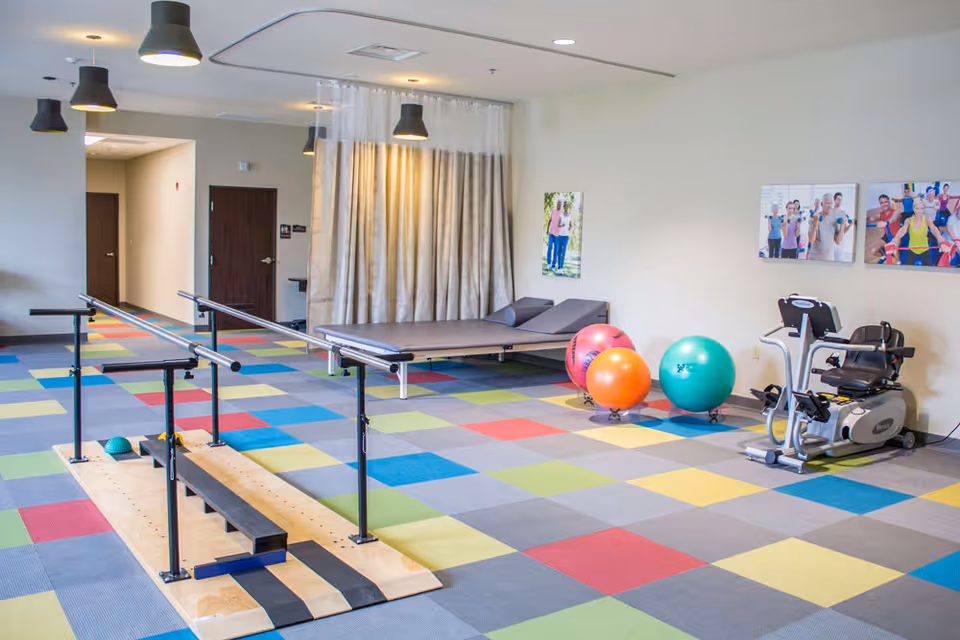 Bright rehabilitation/exercise room with parallel bars, a treatment table, colorful square floor tiles, exercise balls, and cardio equipment.