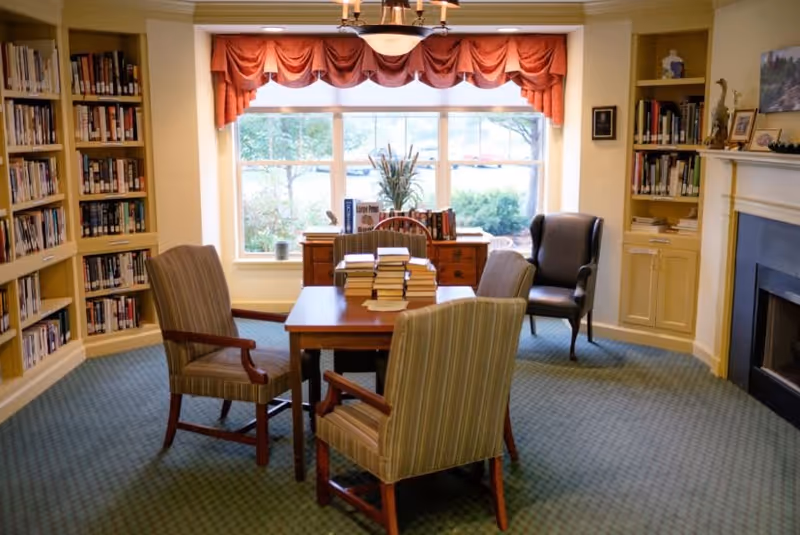 A cozy reading room with a table and four chairs, bookshelves, a fireplace, and a large window with a red valance.