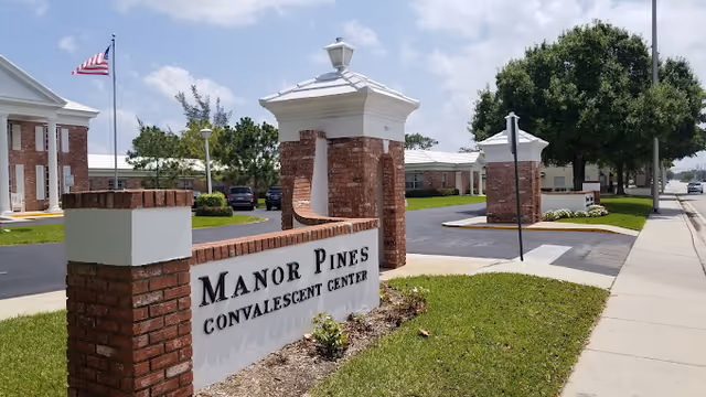 Entrance sign for Manor Pines Convalescent Center made of brick and white panels, with a building and American flag in the background under a partly cloudy sky.