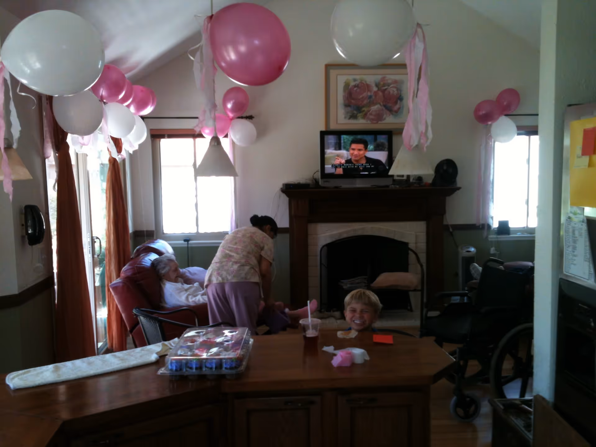 A living room decorated with pink and white balloons and streamers. An elderly person is seated in a recliner chair near a window, while a caregiver tends to them. A young child is smiling at a wooden table in the foreground. A TV is mounted above a fireplace showing a man speaking. There is a wheelchair near the window on the right side.