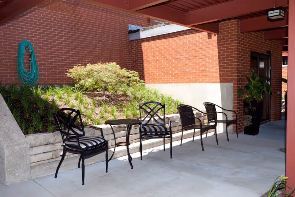Outdoor patio area with four black metal chairs, two with striped cushions, and a small round table between them. The area is adjacent to a red brick wall with some greenery and a garden hose mounted on the wall. There is a large potted plant near a glass door under a red metal beam structure.