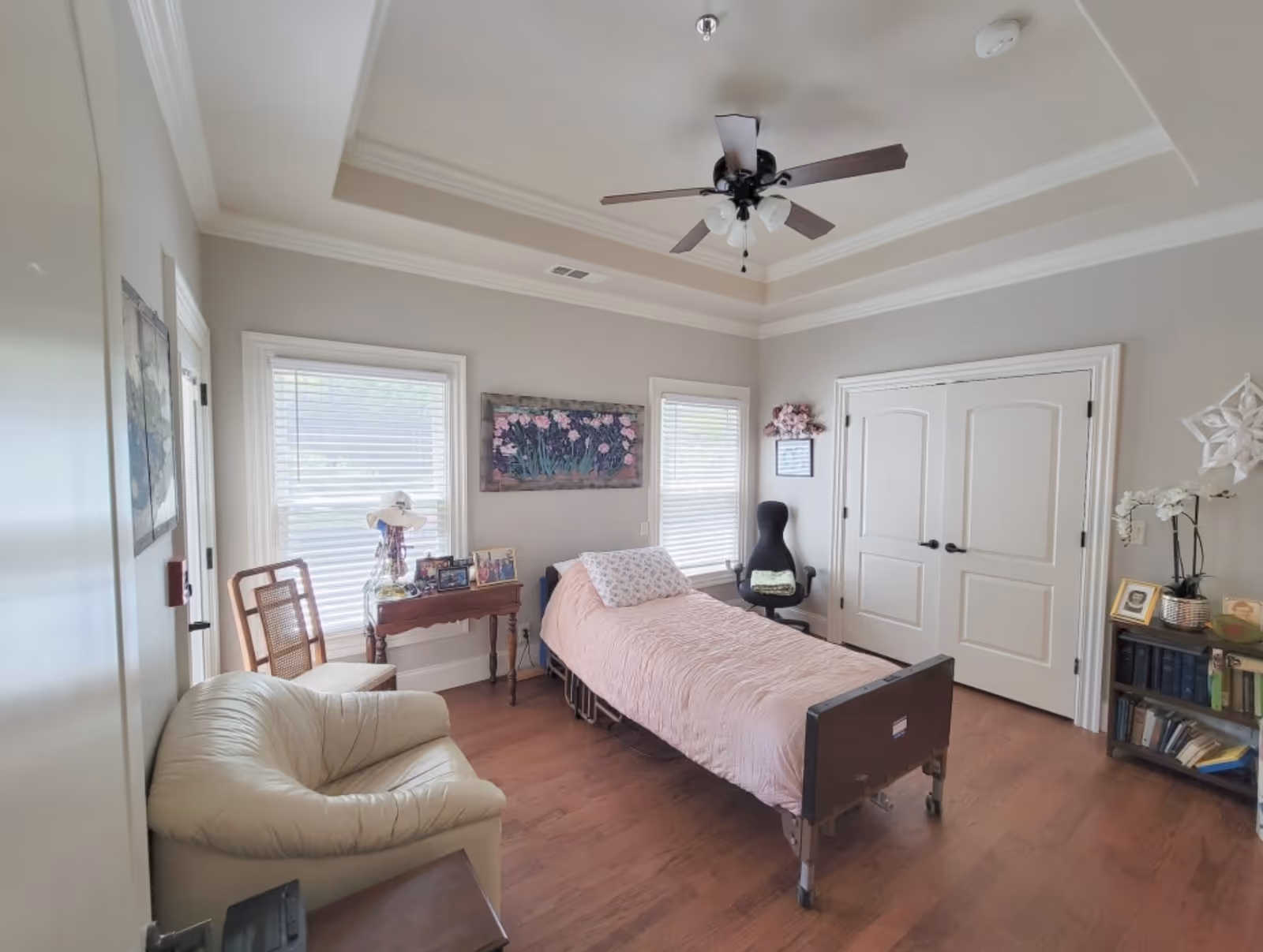 A well-lit bedroom with a single hospital-style bed covered with a pink quilt, a beige armchair, a wooden chair, a small wooden table with framed photos and a hat, a black office chair, and a bookshelf with books and decorative items. The room has two windows with blinds, a ceiling fan with lights, and double white doors.