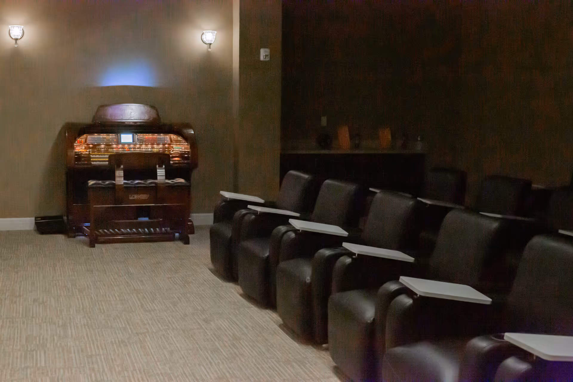 A dimly lit room with a row of black leather chairs equipped with small white foldable desks, facing an antique organ against the wall illuminated by two wall sconces.