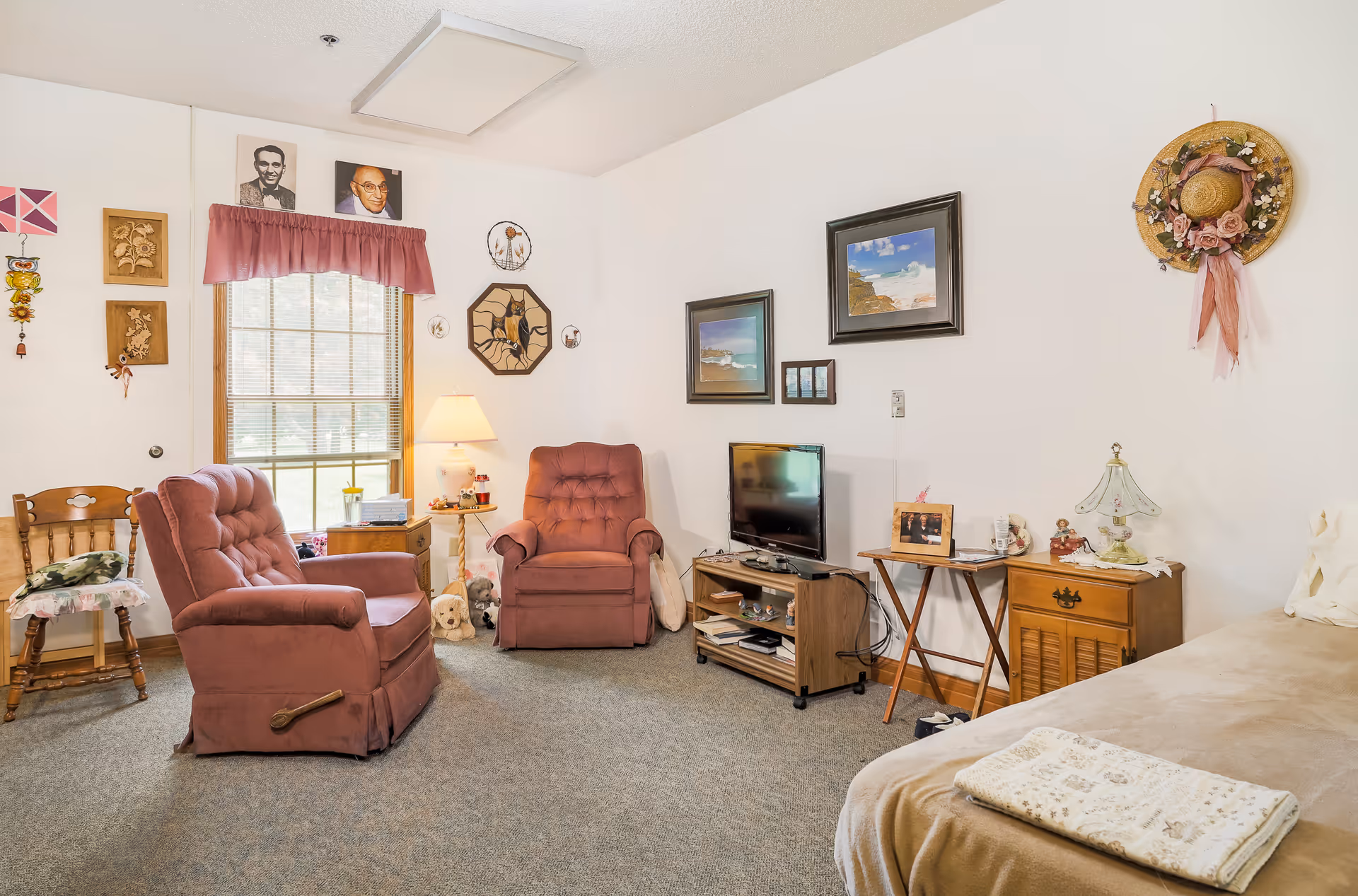 A cozy living room with two pink upholstered armchairs, a wooden chair with a cushion, a small wooden TV stand with a flat-screen TV, and various framed pictures and decorative items on the walls. There is a window with blinds and a pink valance, a side table with a lamp, and a beige couch with a folded blanket on it.