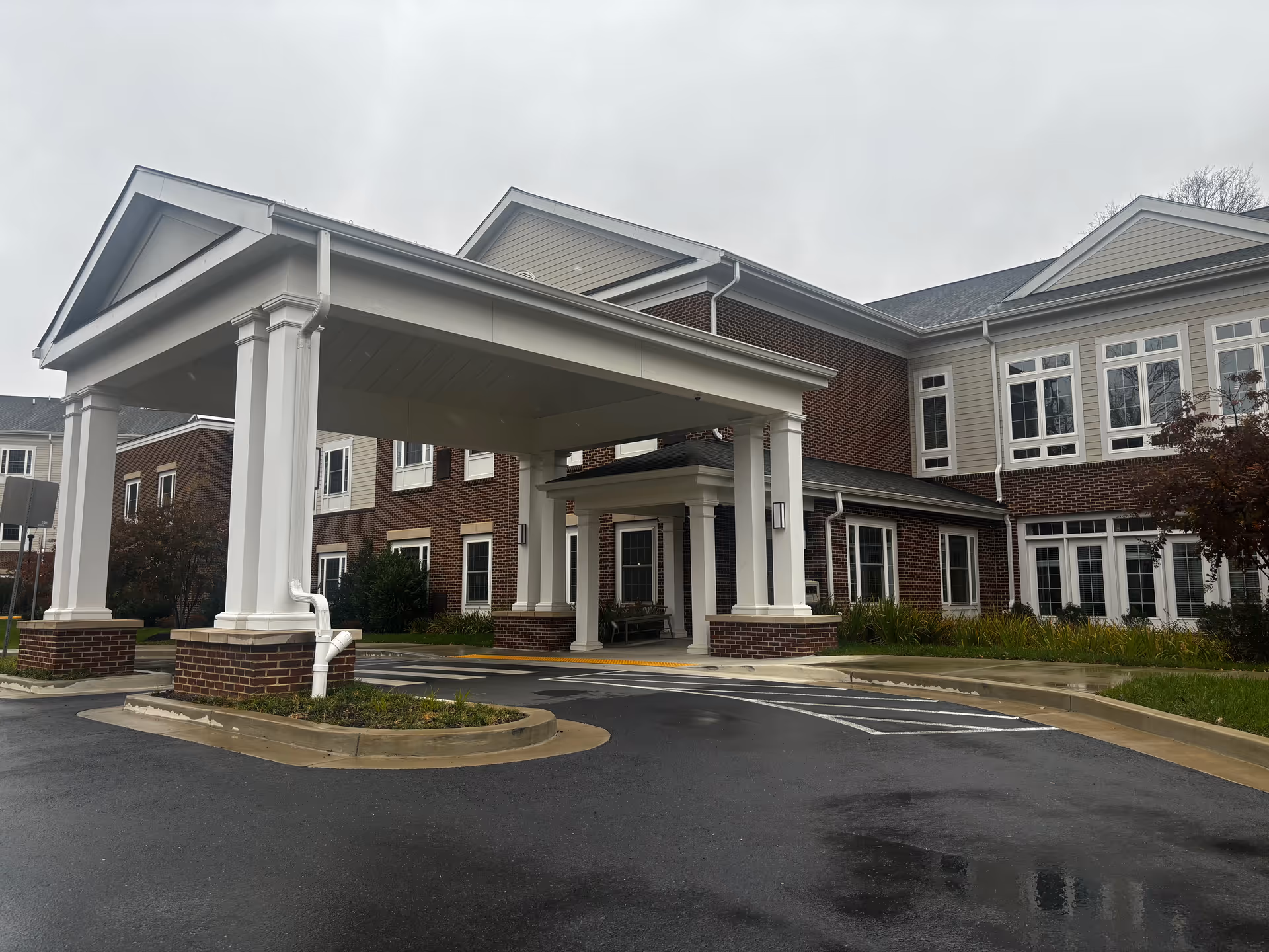 Exterior view of HarborChase of Olney senior living facility showing the main entrance with a covered drop-off area supported by white columns, brick and siding building facade, multiple windows, and a wet driveway on a cloudy day.
