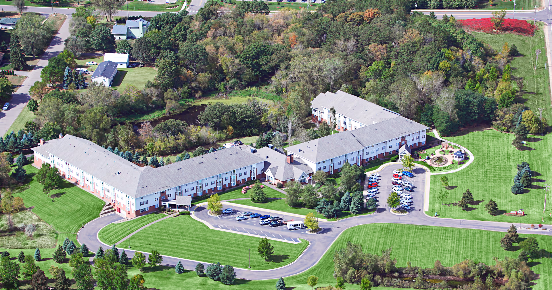 Aerial view of a large senior living facility complex surrounded by green lawns, parking lots, trees and wooded areas.